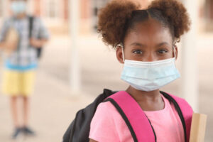 Back to school. African descent girl on school campus. She wears a mask for COVID-19, Coronavirus protection. Other student in background.